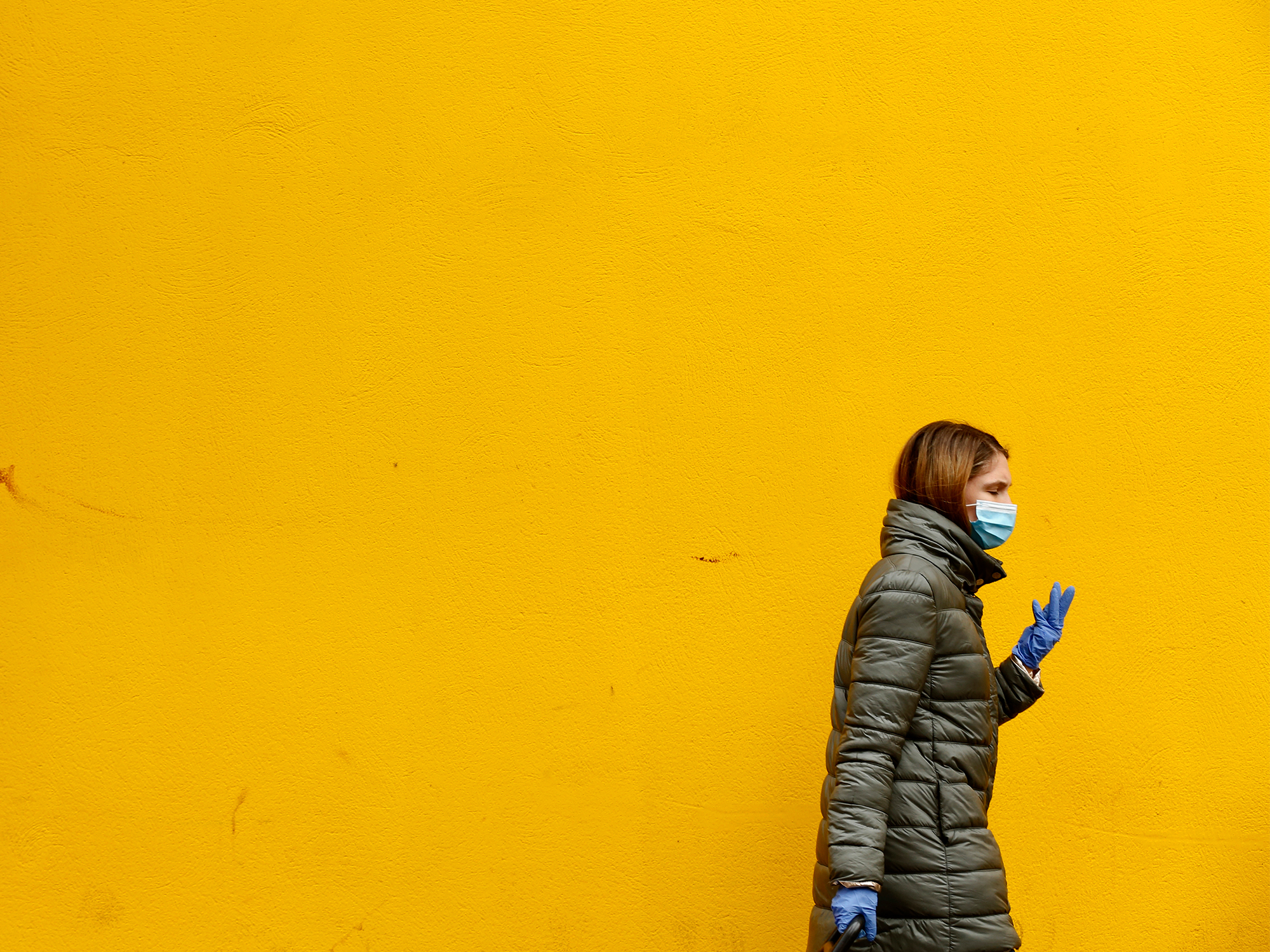Woman walking along a yellow wall while wearing a face mask.